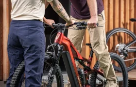 Dos personas están de pie junto a una bicicleta eléctrica de montaña FOCUS e-MTB 2027 de color rojo, sujetando el manillar y el sillín, y en el fondo se ve otra bicicleta, posiblemente un modelo JAM o SAM.