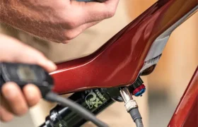 Una persona que sostiene un smartphone cerca de la suspensión trasera de una bicicleta eléctrica de montaña FOCUS e-MTB 2027 de color rojo, y que parece estar comparando datos o realizando ajustes.