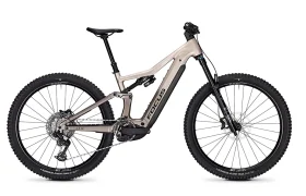 Una bicicleta de montaña eléctrica de doble suspensión FOCUS e-MTB 2027 de color beige con ruedas negras, batería central, frenos de disco y neumáticos anchos con tacos, mostrada de perfil sobre un fondo blanco.