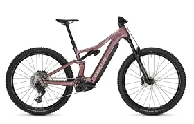 Esta bicicleta eléctrica de montaña FOCUS, de suspensión total y color rosa mate, cuenta con neumáticos de tacos, frenos de disco y un motor de tracción central. La imagen, tomada de perfil sobre un fondo blanco, plasma el espíritu robusto de las series JAM y SAM.