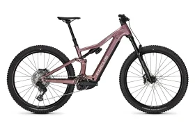 Una bicicleta de montaña de doble suspensión de la gama FOCUS e-MTB 2027, con cuadro rosa, ruedas negras, neumáticos anchos, frenos de disco y un motor central visible, fotografiada de perfil sobre un fondo blanco.