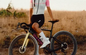 Un ciclista, equipado con casco y ropa de ciclismo, se encuentra junto a una bicicleta Van Rysel EDR-G en un camino de tierra situado en un campo abierto y cubierto de hierba, listo para enfrentarse al terreno de grava.