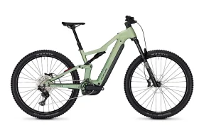 Una bicicleta de montaña eléctrica (e-MTB) FOCUS de color verde claro, con suspensión total, frenos de disco, neumáticos anchos y una batería visible integrada en el cuadro.