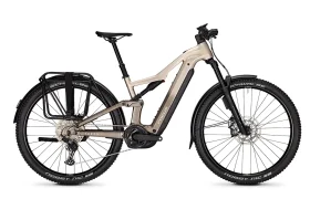 Una bicicleta eléctrica FOCUS e-MTB de color beige con motor central, frenos de disco, portaequipajes trasero, horquilla de suspensión delantera y neumáticos grandes con banda de rodadura.