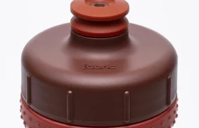 Primer plano de una botella de agua de plástico «Fabric Gripper V2» de color marrón y rojo, con un agarre texturizado y la palabra «fabric» grabada en relieve en el tapón. Esta botella antimicrobiana ofrece protección permanente, lo que la hace ideal para la hidratación diaria.