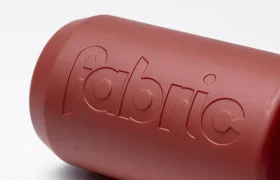 Un primer plano de un recipiente cilíndrico rojo, el Fabric Gripper V2, con la palabra «fabric» grabada en relieve en uno de sus laterales, sobre un fondo blanco liso. Este bidón antimicrobiano está diseñado para ofrecer una protección permanente.