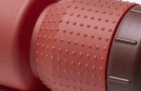 Primer plano de un agarre de goma rojo con textura —conocido como «Fabric Gripper V2»— en un bidón cilíndrico con tapón de color marrón oscuro, sobre un fondo blanco. Este diseño incorpora protección antimicrobiana para una mayor higiene.