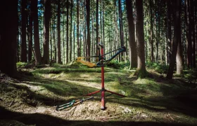 Un cuadro de bicicleta de montaña, con la horquilla FOX 38 Racing Green, está montado en un puesto de reparación en un bosque iluminado por el sol, con herramientas y piezas de bicicleta esparcidas por el musgoso suelo cercano.