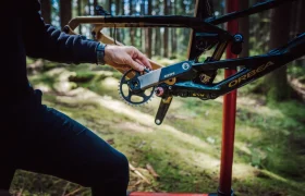 Una persona ajusta los platos y bielas de una bicicleta Orbea con horquilla FOX 38 Racing Green, montada sobre un caballete en un bosque iluminado por el sol.