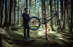 Una persona ajusta una bicicleta de montaña con un FOX 38 Racing Green Edición Limitada en un puesto de reparación en un bosque, rodeado de altos árboles y la luz del sol filtrándose a través del dosel.