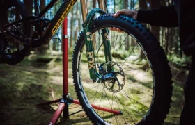 Una persona instala una rueda delantera en una bicicleta de montaña equipada con una suspensión FOX 38 Racing Green Edición Limitada en una zona boscosa, utilizando un soporte de bicicleta rojo como apoyo.
