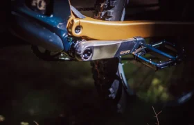 Primer plano de un brazo de biela de bicicleta con la etiqueta "EXR" y pedal plano fijado al cuadro naranja y azul, acentuado por la horquilla FOX 38 Racing Green, con parte de la rueda trasera y la cadena visibles.