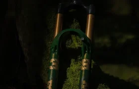 Una horquilla de suspensión de bicicleta de montaña FOX 38 Racing Green está apoyada contra un árbol musgoso en un bosque poco iluminado, mostrando su llamativo acabado verde y dorado Edición Limitada.