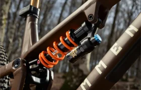 Primer plano de un amortiguador trasero FOX de bobina naranja en una bicicleta de montaña de suspensión integral marrón, mostrando la nueva generación de rendimiento con un fondo de bosque difuminado.