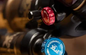 Primer plano de una horquilla de suspensión de bicicleta de montaña con mandos de ajuste rojos y azules etiquetados como "Rebound" (rebote) y "Firm/Low Speed Compression" (firme/baja velocidad de compresión), mostrando el control y ajuste de amortiguadores FOX.