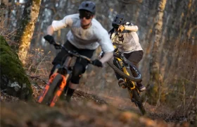 Dos ciclistas de montaña bajan por un sendero forestal, uno de ellos levanta la rueda delantera mientras sigue de cerca al otro, mostrando el control y el suave rendimiento de los amortiguadores FOX.