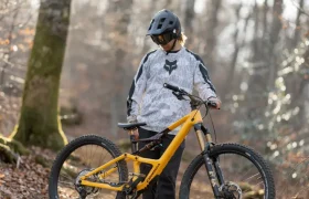 Una persona con casco y ropa de ciclista se encuentra en un sendero de tierra en el bosque, sujetando una bicicleta de montaña amarilla equipada con amortiguadores de primera calidad.