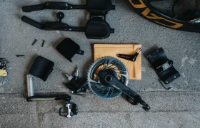 Los componentes de la bicicleta, que incluyen platos y bielas, pedales, extensiones de manillar y varios tornillos, están meticulosamente dispuestos sobre una superficie de hormigón. Un potenciómetro de Body Rocket añade un toque avanzado a la configuración.