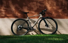 Una moderna bicicleta de carretera Van Rysel RCR-G, con manillar de carretera y neumáticos finos, se apoya contra una pared texturizada en dos tonos, proyectando una sombra sobre el césped que hay debajo; lista tanto para el asfalto liso como para explorar por caminos de grava suelta.