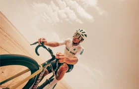 Un ciclista, vestido con ropa blanca y casco, pedalea en una elegante bicicleta de carretera Van Rysel RCR-G por una carretera abierta y soleada, con un cielo despejado al fondo.