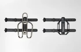 Dos soportes de portabidón para bicicleta con correas se muestran uno junto al otro sobre un fondo liso; el de la izquierda sostiene un portabidón metálico, mientras que el de la derecha presenta un portabidón de plástico negro, ambos compatibles con los sistemas Tailfin Hydromount.