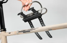 Una mano instala un portabidón metálico Tailfin Hydromount con dos correas en el tubo superior del cuadro de una bicicleta beige.