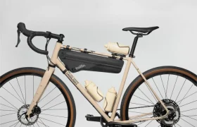 Una bicicleta de grava beige con manillar negro, una bolsa de cuadro y tres bidones de agua beige montados en el cuadro con soportes hidráulicos Tailfin, sobre un fondo liso.