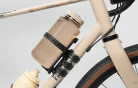 Un cuadro de bicicleta beige con una gran botella de agua transparente sujeta en un Tailfin Hydromount negro y una botella amarilla más pequeña en un segundo soporte.