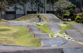 Un ciclista recorre una pista de pump track pavimentada con múltiples baches y curvas, equipada con una tija telescópica inalámbrica, rodeado de césped, árboles y un edificio al fondo.
