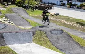 Una persona con casco monta en una bicicleta BMX con tija telescópica inalámbrica RAGE Circuit, cogiendo aire sobre un salto de tierra y asfalto en una pista de pump track al aire libre.