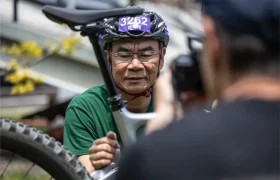 Un hombre con casco y gafas inspecciona una bicicleta equipada con una tija telescópica inalámbrica RAGE Circuit, mientras otra persona en primer plano hace una foto o graba un vídeo.