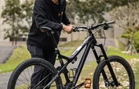 Un hombre vestido de negro está de pie al aire libre sobre la hierba, sujetando una bicicleta de montaña negra con pedales naranjas y una tija telescópica inalámbrica RAGE Circuit. Al fondo se ven árboles y un edificio.