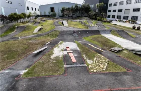 Una pista de pump track al aire libre con caminos pavimentados, zonas de hierba, rampas y obstáculos es perfecta para poner a prueba tu tija telescópica inalámbrica Circuito RAGE, todo ello en medio de naves industriales y algunos árboles.