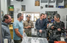 Seis hombres permanecen juntos en un taller o laboratorio, inmersos en una conversación; uno de ellos, a la derecha, sostiene y gesticula con una tija telescópica inalámbrica RAGE Circuit.