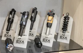 Cinco amortiguadores de bicicleta EXA Form se muestran en posición vertical sobre soportes, con dos muelles adicionales sobre el estante de cristal, perfectos junto a su configuración de tija telescópica inalámbrica RAGE Circuit o tija telescópica KS.