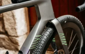 Primer plano de una bicicleta Ridley Kanzo Fast 2.0 con una pequeña bolsa negra de Ridley sujeta al manillar, que presenta detalles de diseño aerodinámico y neumáticos anchos de 58 mm, aparcada junto a un muro de piedra y una puerta de madera.