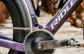 Primer plano de la transmisión de una bicicleta Ridley Kanzo Fast 2.0, en el que se aprecian el juego de bielas, la cadena, los pedales y parte del cuadro aerodinámico con dos portabidones vacíos. La bicicleta está aparcada sobre una superficie de piedra, lista para rodar con sus robustos neumáticos de 58 mm.