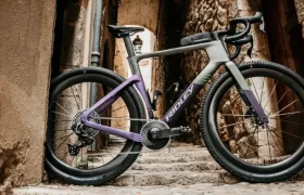 Una moderna bicicleta de carretera, la Ridley Kanzo Fast 2.0, con un cuadro morado y gris y características aerodinámicas, está aparcada en unos escalones de piedra entre antiguos y estrechos edificios de piedra en un callejón.