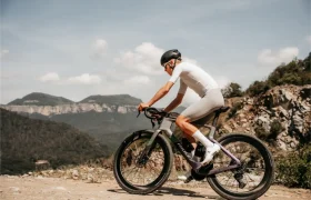 Una persona que lleva casco y equipación de ciclismo circula en una bicicleta Ridley Kanzo Fast 2.0, equipada con características aerodinámicas y neumáticos de 58 mm, por un camino de grava, con montañas y terreno rocoso al fondo, bajo un cielo parcialmente nublado.