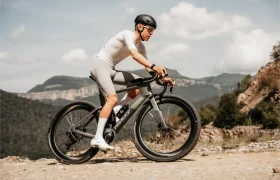 Un ciclista, con casco y equipación de ciclismo, circula en una bicicleta de gravel Ridley Kanzo Fast 2.0 con neumáticos de 58 mm por un sendero rocoso, con montañas y árboles al fondo.