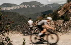 Dos ciclistas en una ruta de montaña; uno está de pie junto a una Ridley Kanzo Fast 2.0 —con su cuadro aerodinámico y sus neumáticos de 58 mm, preparados para cualquier terreno—, de cara a unos acantilados lejanos, mientras que el otro pasa en bicicleta por el primer plano, difuminado por el movimiento.