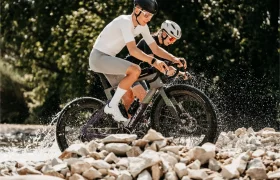 Dos ciclistas, con casco y gafas de sol, pedalean en unas bicicletas de gravel Ridley Kanzo Fast 2.0, equipadas con manillares aerodinámicos y robustos neumáticos de 58 mm, a través de un arroyo poco profundo y rocoso, salpicando agua mientras pedalean. Al fondo se ven árboles y vegetación.