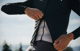 Una persona se abrocha una chaqueta negra ASSOS EQUIPE sobre una camisa blanca al aire libre; se le ve parte del torso y las manos sobre un fondo difuminado, lo que destaca la protección ultraligera y las innovadoras características del ThermoBooster.