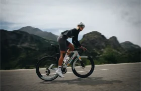 Una persona que lleva casco y equipación ciclista, incluida una chaqueta ASSOS EQUIPE ThermoBooster para una protección ultraligera durante el entretiempo, circula en bicicleta de carretera por una carretera asfaltada con montañas al fondo, bajo un cielo nublado.
