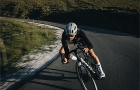 Un ciclista que lleva casco y gafas de sol se inclina al tomar una curva en una carretera asfaltada, con colinas cubiertas de hierba al fondo, confiando plenamente en el ASSOS EQUIPE ThermoBooster para disfrutar de una protección ultraligera durante esos días impredecibles de entretiempo.
