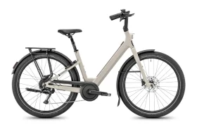 Una bicicleta eléctrica Moustache Lundi 27 de color beige, con cuadro abierto, guardabarros negros, portaequipajes trasero y neumáticos anchos, mostrada de perfil sobre un fondo blanco: perfecta para desplazarse con comodidad por la ciudad.
