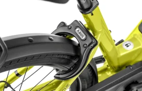 Primer plano de un candado de anillo negro de la marca ABUS fijado a la rueda trasera y al cuadro de una bicicleta amarilla Moustache Lundi 27, perfecta para moverse por la ciudad con total seguridad.