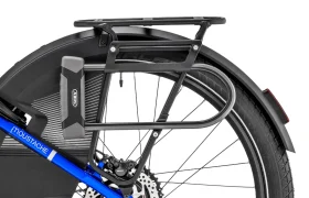 Primer plano de la rueda trasera de una bicicleta azul «Moustache Lundi 27», en el que se aprecian un portaequipajes trasero negro, un guardabarros, un freno de disco, un candado ABUS y parte del neumático: perfecta para moverse por la ciudad.