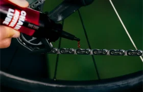 Una mano aplica lubricante para cadenas a la cadena de una bicicleta, y una gota de lubricante cae sobre los eslabones metálicos cerca de la rueda trasera.