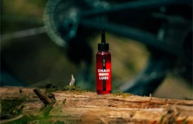 Una botella roja de lubricante para cadenas Motul está colocada en posición vertical sobre un tronco al aire libre, con una rueda de bicicleta borrosa visible al fondo.